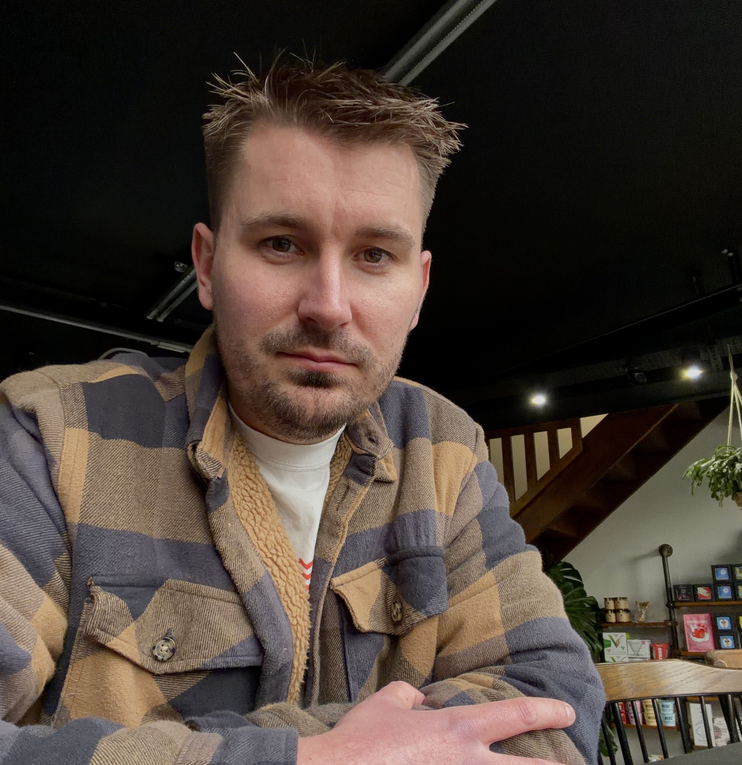 A selfie in a cafe. I'm wearing a white t-shirt with a blue and tan lumberjack over the top. I'm looking into the camera with a soft smile
